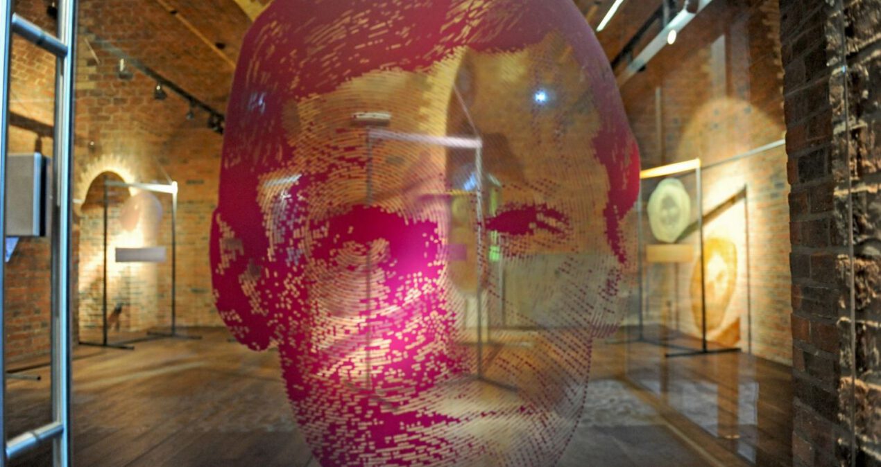 Na pierwszym planie różowa grafika prezentująca twarz mężczyzny. Umieszczona jest na przezroczystym tle, przez które widać wnętrze sali wystawowej